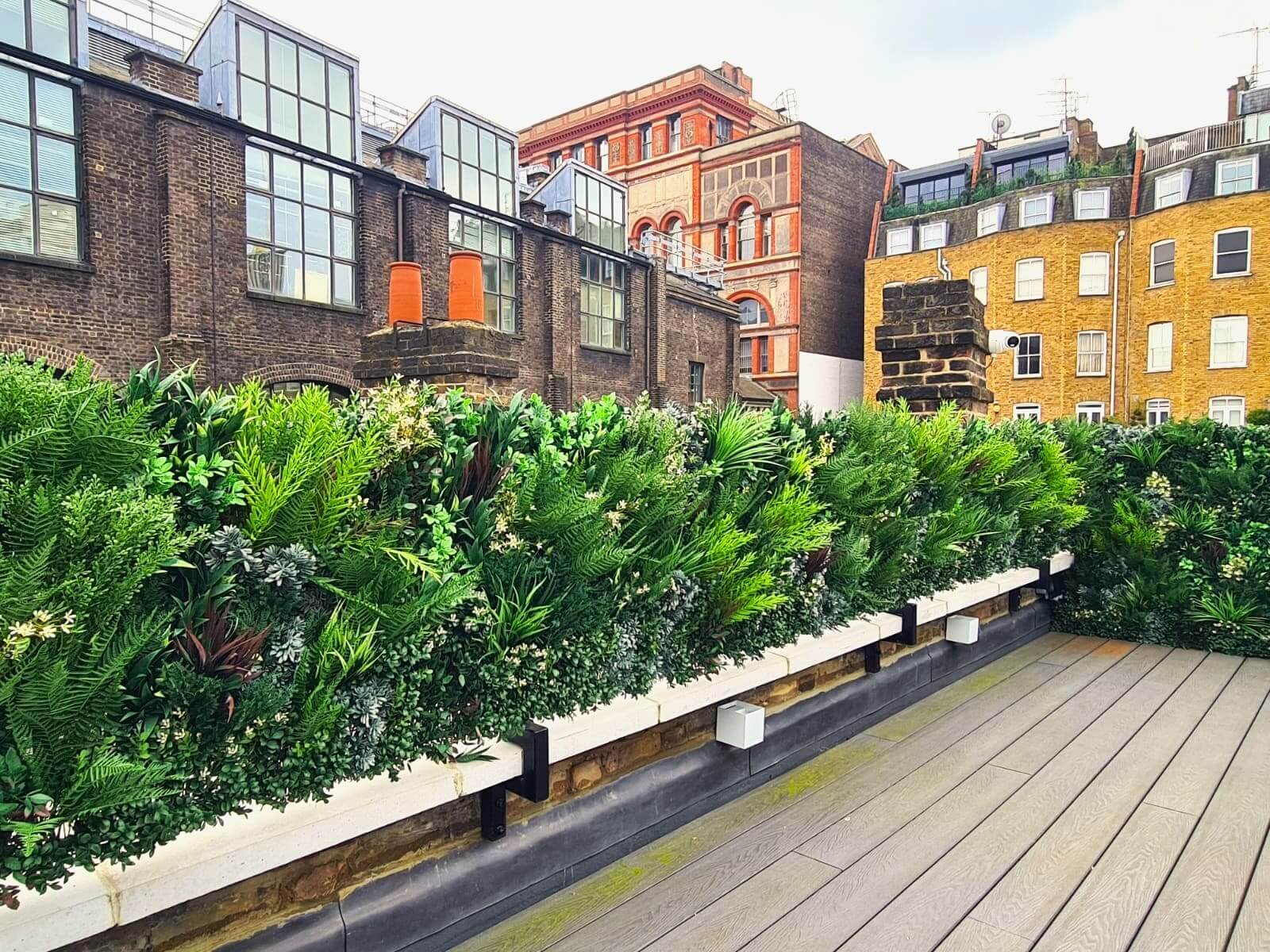 A rooftop garden with lush green plants and flowers along a wooden deck, set against a backdrop of historic brick buildings with large windows. The sky is overcast, creating a striking contrast between the greenery and the urban architecture enhanced by artificial touches.