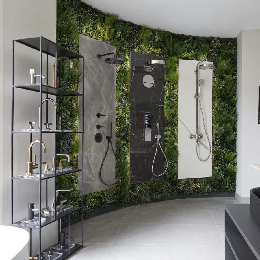 An artificial green wall provides a striking backdrop in this modern bathroom showroom, showcasing three unique shower setups with diverse showerheads and controls. A sleek black shelving unit to the left displays an assortment of faucets and bathroom fixtures.