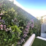 A lush vertical garden with various green plants and purple flowers, a bespoke transformation with Vistafolia, covers an outdoor wall. A staircase is visible on the right, and sunlight brightens the scene under a clear blue sky, unleashing imagination in this serene setting.