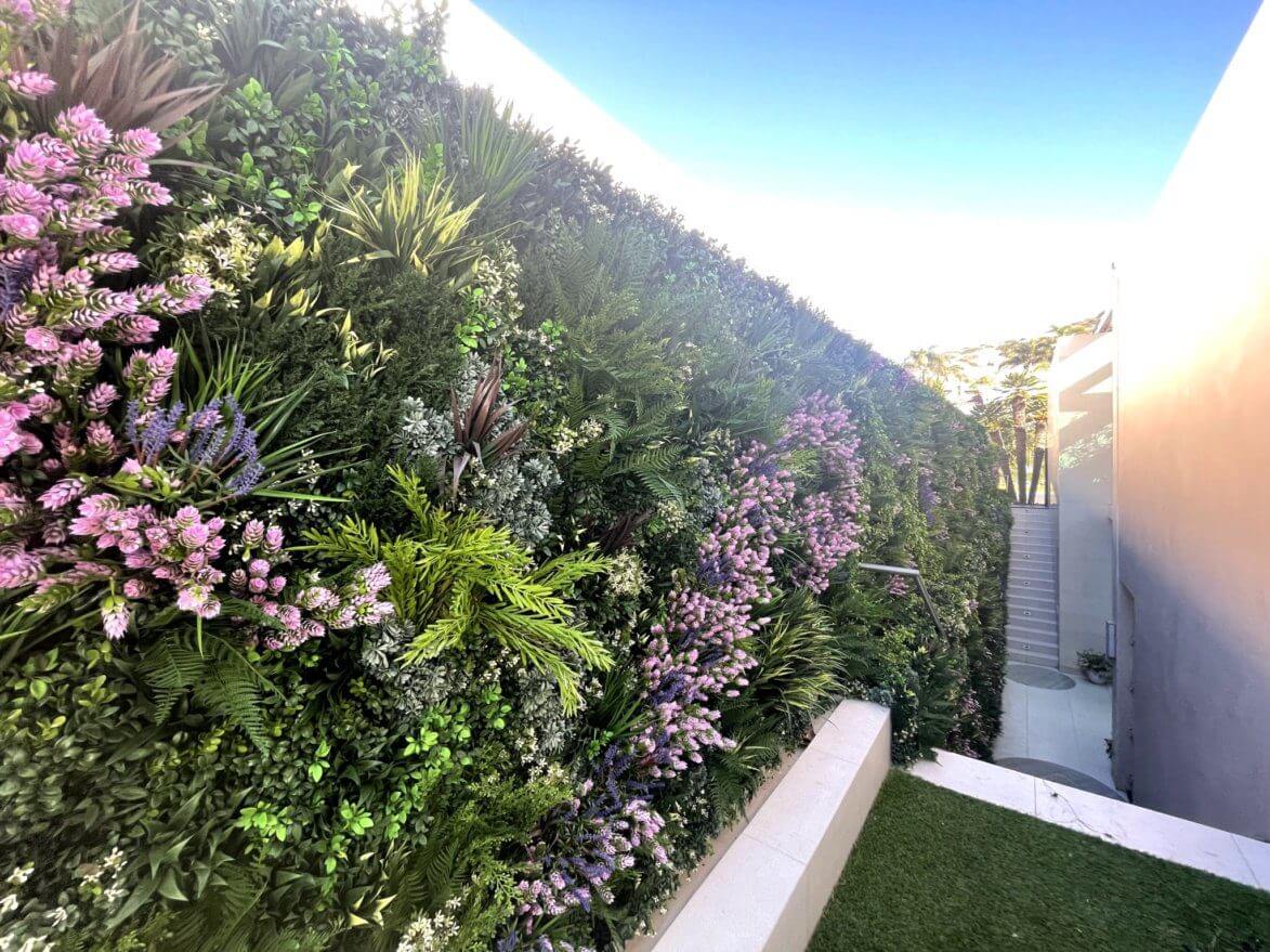 An artificial vertical garden, vibrant with green plants and purple flowers, covers a tall wall. A staircase with a white railing is visible on the right side, ascending against a backdrop of clear blue sky.