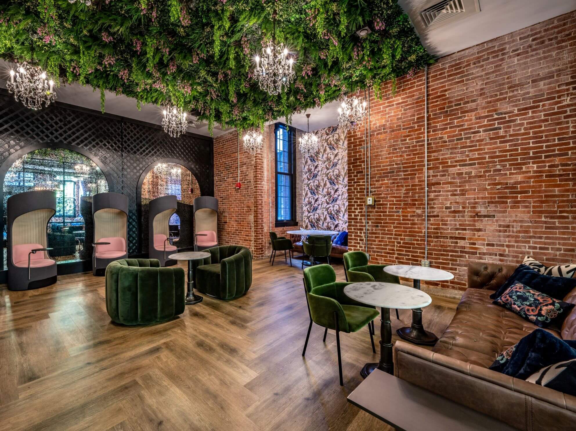 A stylish room in the heart of Rhode Island boasts a brick wall adorned with lush greenery hanging from the ceiling and elegant chandeliers. Featuring velvet chairs, sofas, and small round tables, this chic lounge atmosphere is enhanced by the innovative green ceiling installation in Providence.