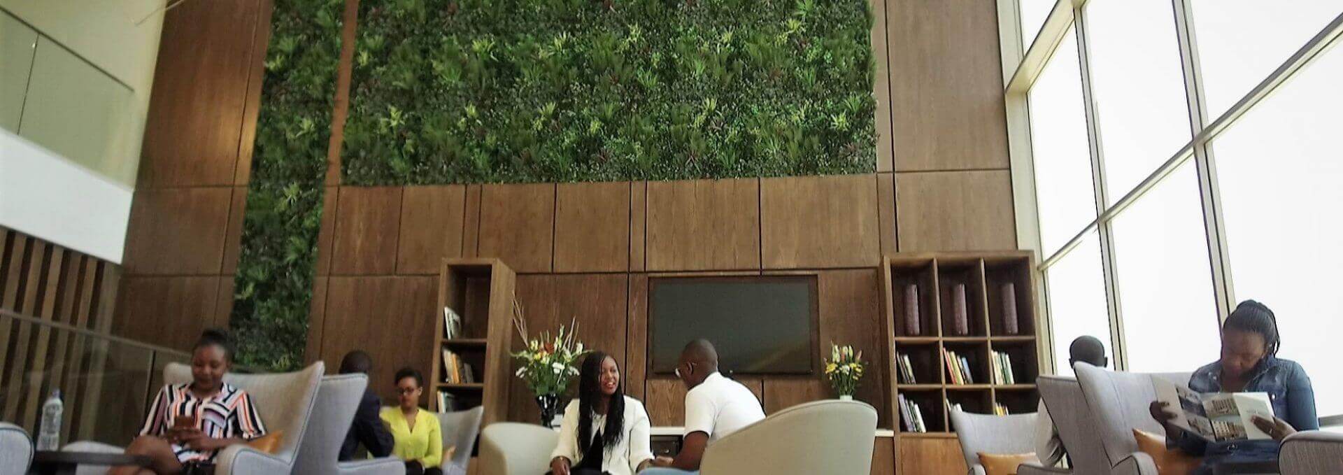 A spacious modern lounge in Nairobi Airport features an artificial green wall and large windows. People are seated in comfortable chairs, some reading and others conversing. Wooden shelves with books and plants decorate the space, while natural light fills the room in Kenya's vibrant hub.