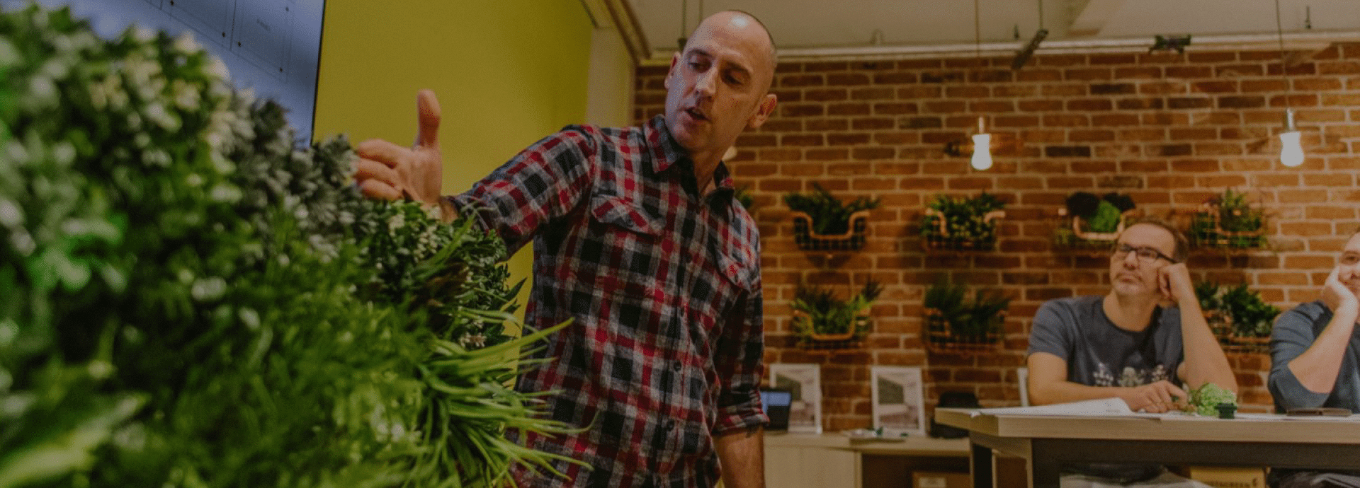 A person in a plaid shirt gestures towards a vertical garden inside an indoor space with brick walls. Another person sits at a table in the background, listening attentively.