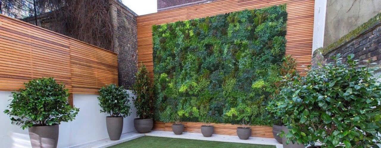 A courtyard featuring a vibrant vertical green wall with lush plants is encased by modern wooden paneling. Several potted plants add to the atmosphere, while the ground is covered with green artificial turf for a fresh look.