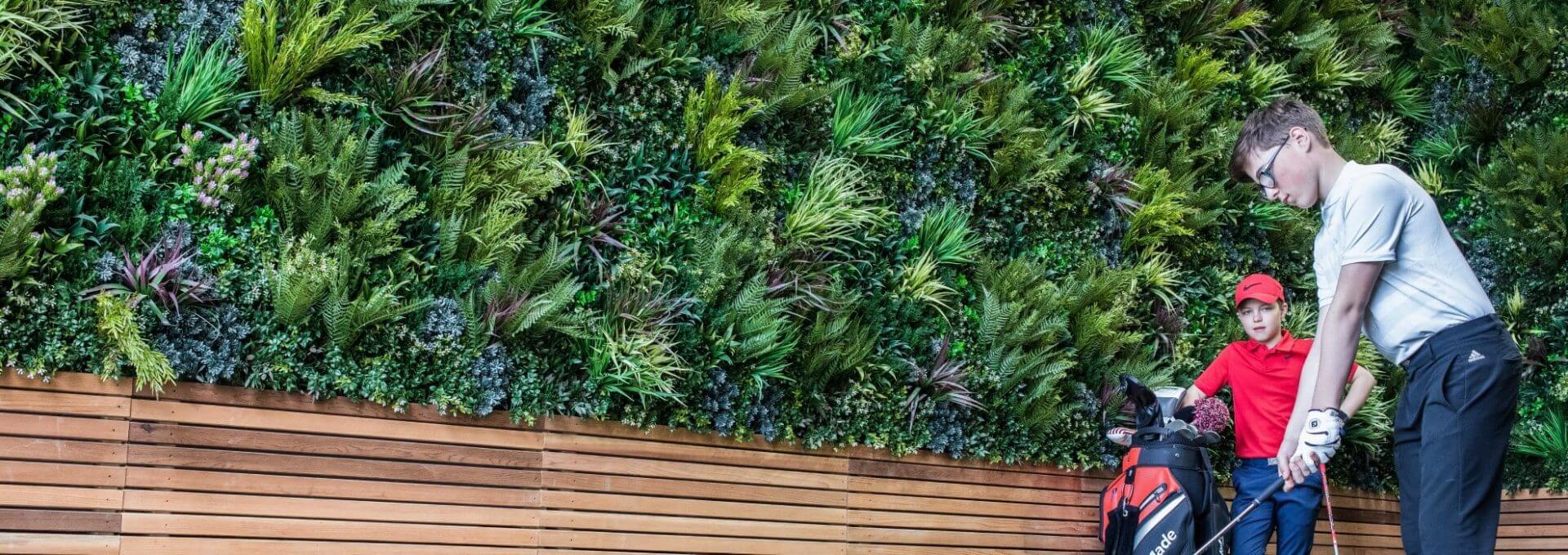 Two young golfers stand indoors; one holds a club while the other leans on a golf bag. An artificial green wall foliage, reminiscent of Portland's natural wonders, covers the wall behind them, and a wooden bench runs along the base.