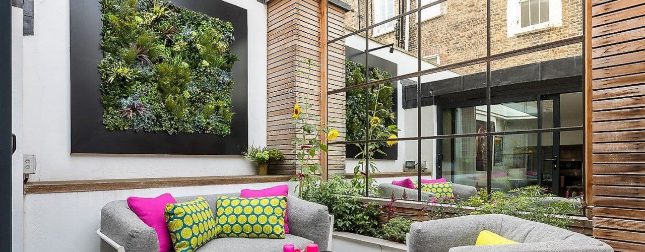 A modern outdoor patio with gray sofas and colorful cushions in pink and green. An artificial vertical garden and large mirror adorn the brick and wood walls, enhancing the vibrant plants and cozy decor.