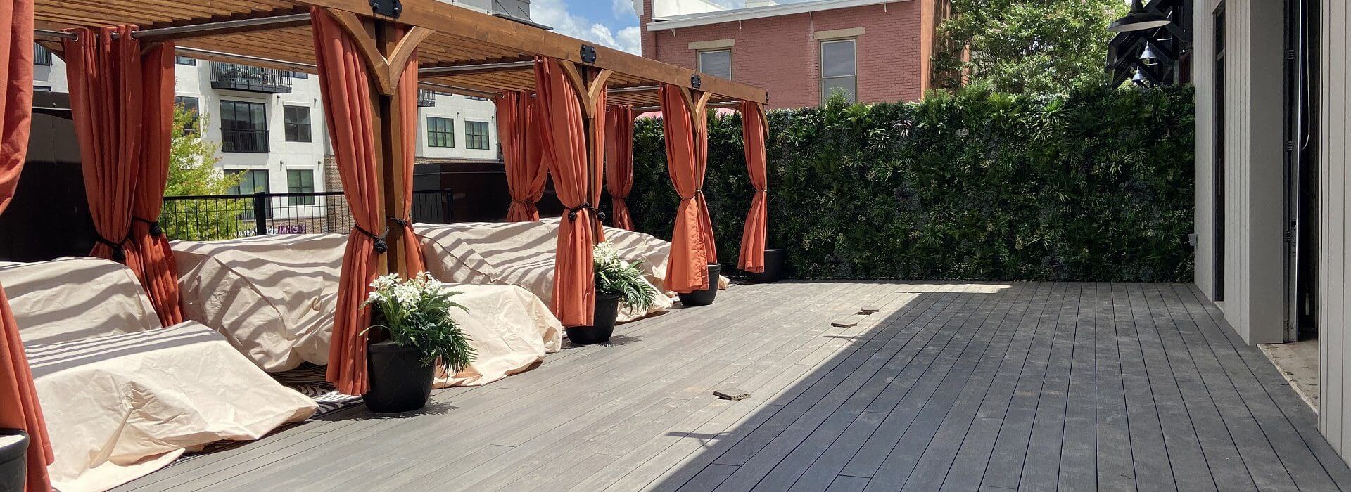 A rooftop terrace with wooden decking features multiple cabanas draped with red curtains, each offering covered seating underneath. Potted plants adorn the space, while a vibrant green wall adds charm to this Huntsville rooftop bar, creating an inviting oasis in Alabama.