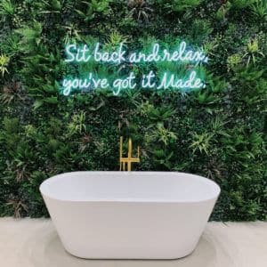 A modern white bathtub is positioned against a lush green plant wall. Above the tub is a neon sign that reads Sit back and relax, you’ve got it Made. A sleek gold faucet is installed on the edge of the tub, giving an artificial touch of elegance to the luxurious setting.