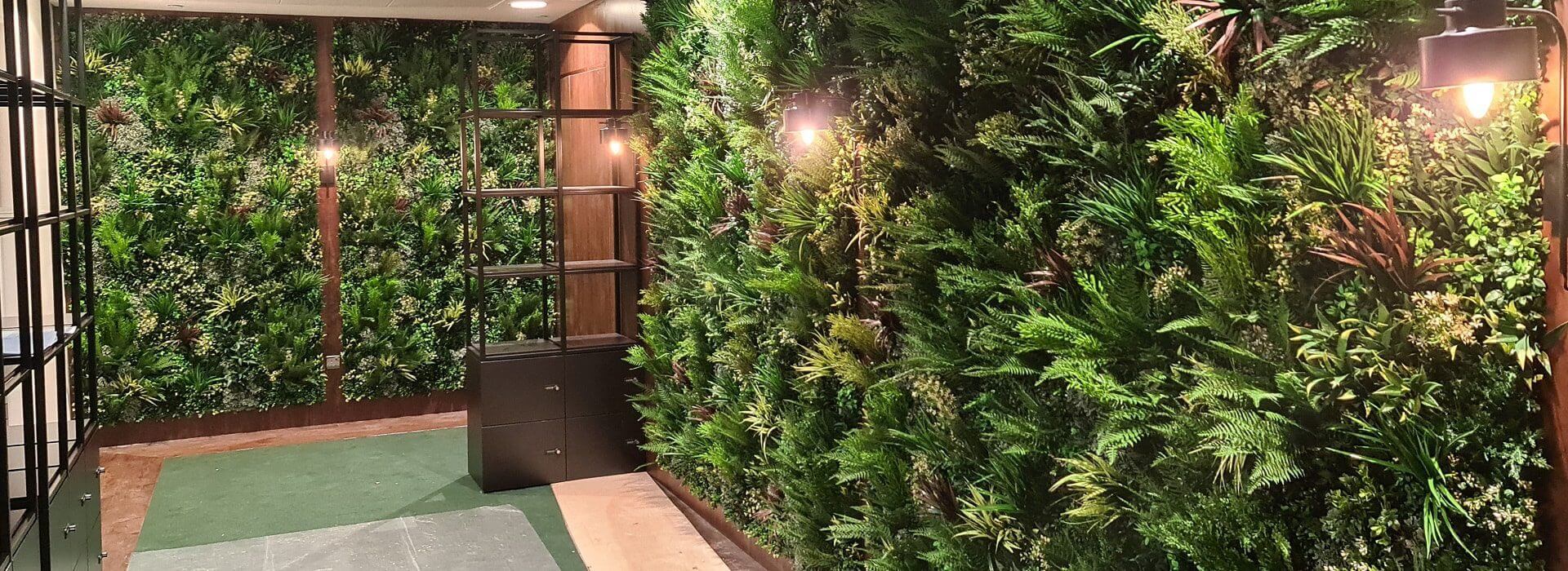 A room in London, UK, boasts three walls draped in an office faux green wall, creating a vertical garden effect. Warm lighting enhances the ambiance while a shelving unit stands on the left, complemented by a plush green carpet underfoot.