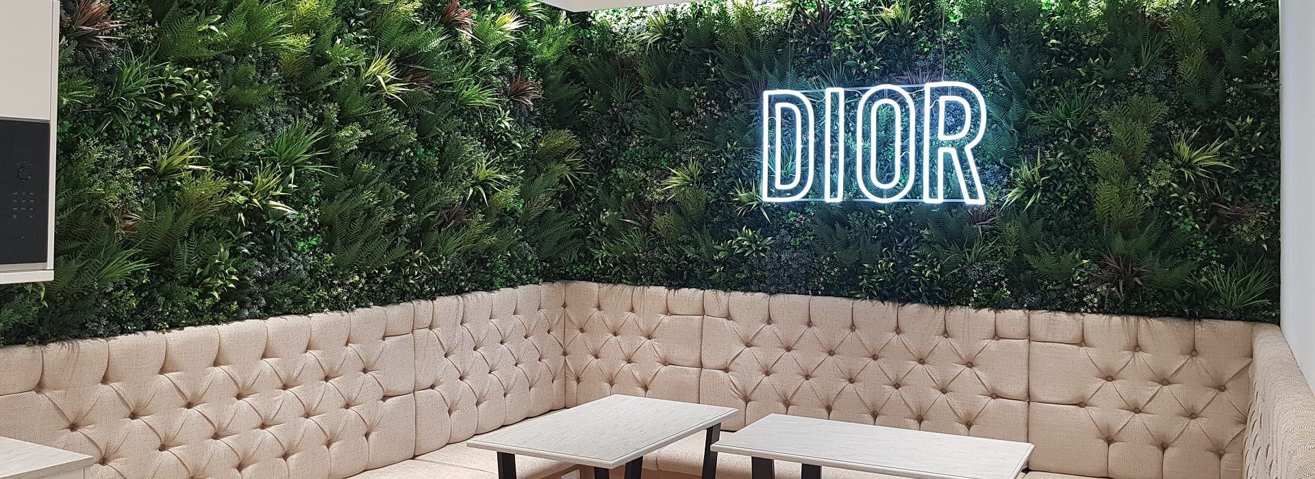 A seating area with beige upholstered benches and light wooden tables is set against a wall covered in lush greenery at the Christian Dior Flagship Store in London. A neon sign reading DIOR illuminates the vibrant green wall, blending elegance with natural charm.