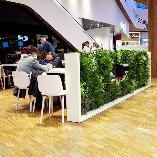 People sit at tables next to a lush green privacy screening in a modern indoor space with wooden flooring. An escalator and various storefronts highlight the vibrant atmosphere of this shopping mall in Milan, Italy.