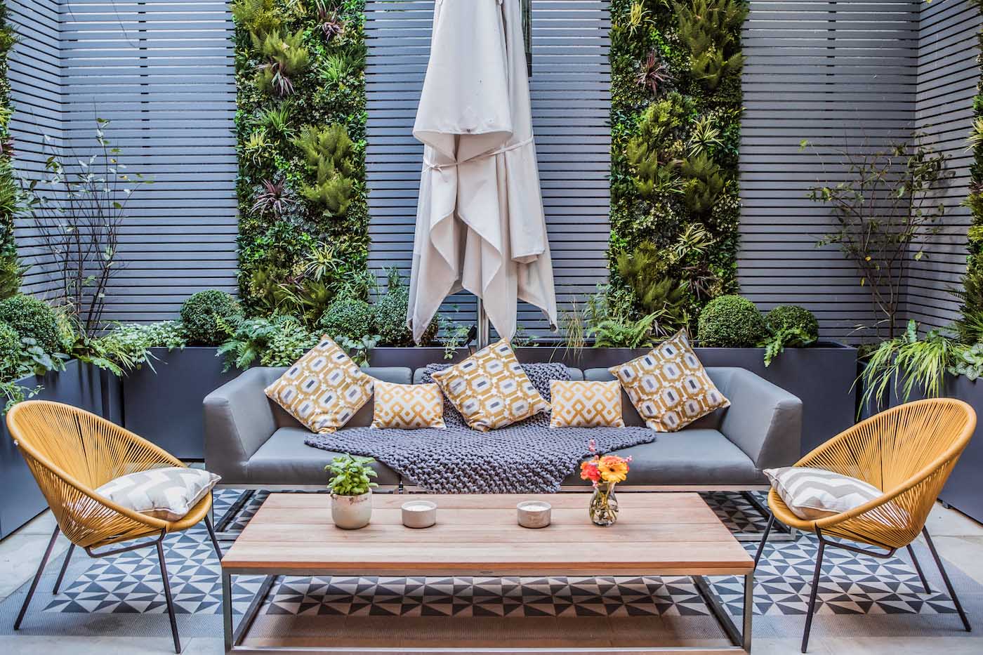 Modern outdoor patio with a gray sofa, patterned pillows, and a gray throw blanket. Two yellow woven chairs flank a wooden coffee table with potted plants and candles. Vertical garden walls and a closed umbrella complete the tranquil setting.