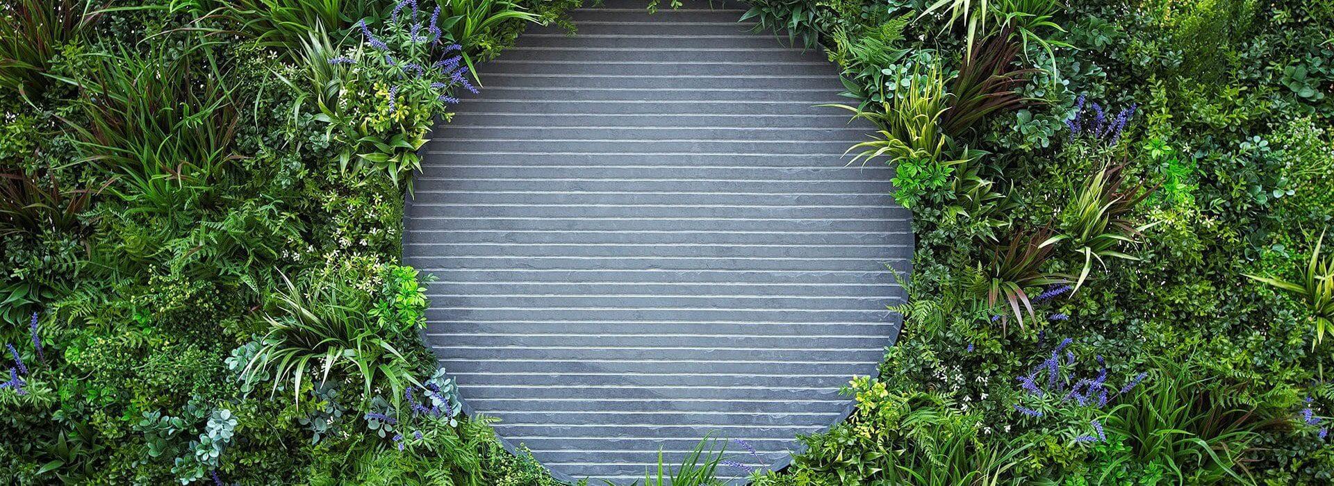 A circular stone wall feature is surrounded by lush green plants and purple flowers, creating an artificial yet natural frame. The foliage showcases various shades of green and textures, providing a vibrant and serene appearance.