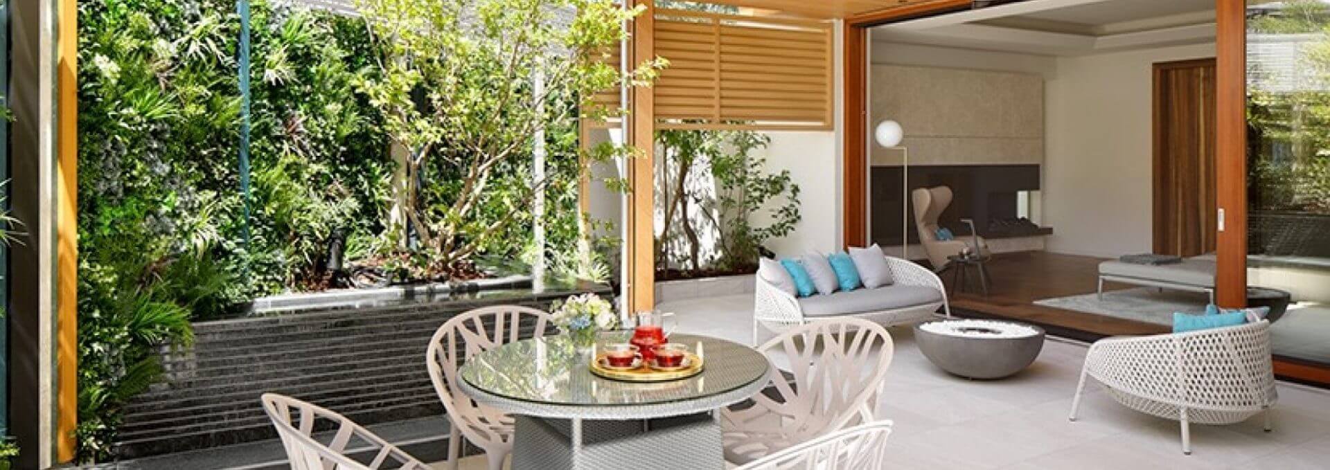 A modern outdoor patio with a round glass table and four chairs, reminiscent of contemporary Japan. The space features a small garden with lush greenery, including a Faux Green Wall in Tokyo style, cushioned seating, and sliding doors leading to an indoor living room.