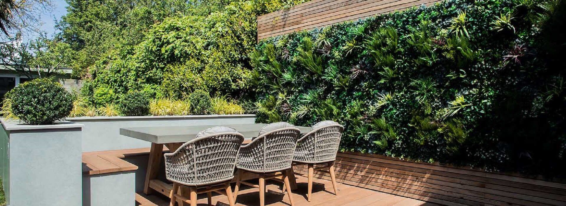 A modern outdoor patio in London, UK, showcases a sleek dining table and four wicker chairs. The space features a lush, artificial green wall set against tall trees and foliage. Manicured shrubs enhance the minimalist design, creating a serene garden oasis in the bustling city.