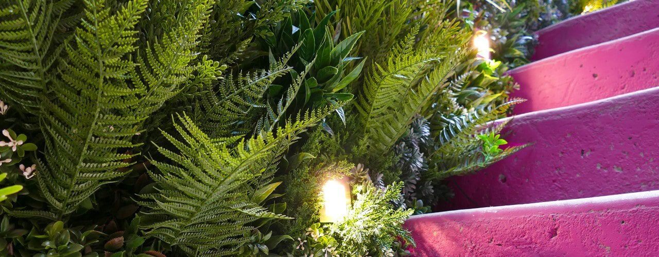 A vibrant staircase painted pink is flanked by lush green ferns and plants. Like a scene from a Vistafolia Guide to Fire Classifications, small lights are interspersed among the foliage, casting a warm glow on the leaves and steps.