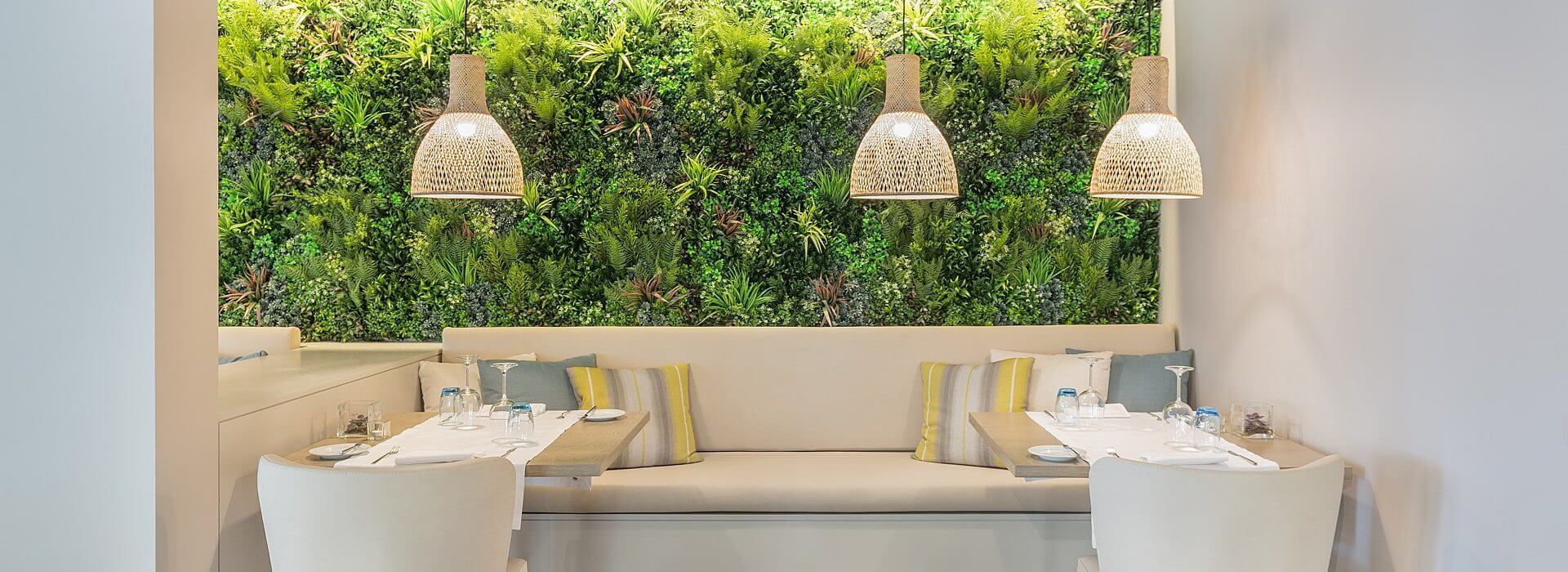 A modern dining area in Lisbon features a bench lined with pillows, two tables set for a meal, and overhead woven lamps. The wall behind is adorned with a lush green wall, offering a fresh and natural ambiance that embodies Portugal's essence of restaurant interior design.