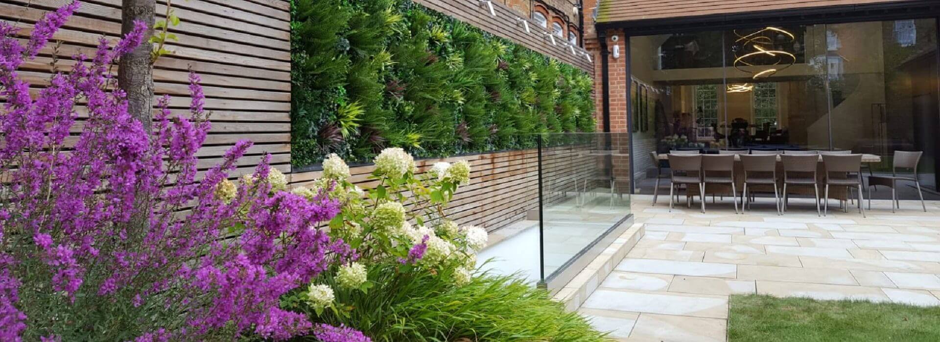 A modern outdoor patio in London boasts garden green walls on wooden panels, adorned with lush plants and vibrant purple flowers. A glass railing separates the patio from a dining area with a large table and chairs inside, framed by floor-to-ceiling glass doors.