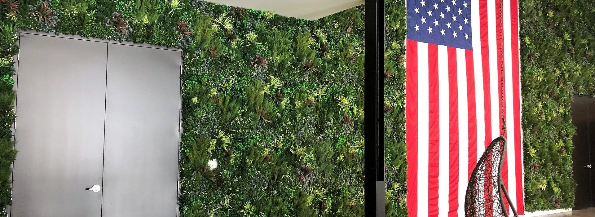 A large American flag hangs vertically on a wall covered with green foliage, giving the room a natural and patriotic theme. A closed black door is on the left, while an artificial net stands in front of the flag, blending seamlessly into the decor.