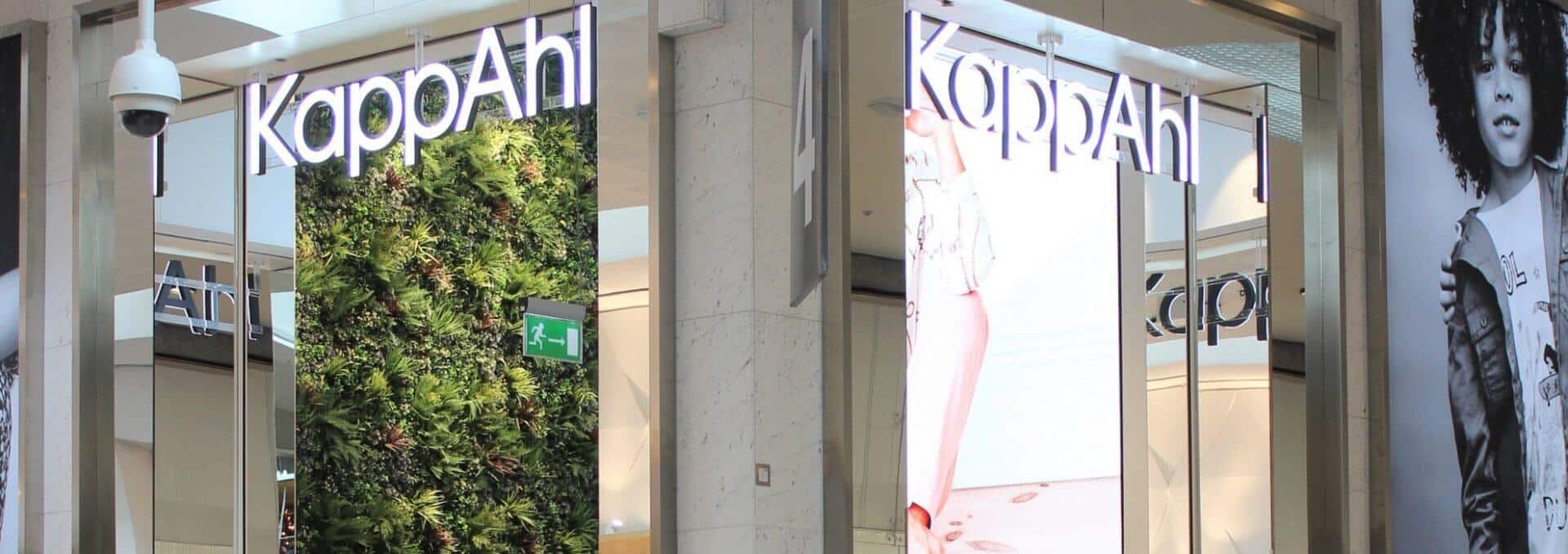 The storefront of KappAhl in Gothenburg features illuminated signage and a striking vertical green wall. Large fashion advertisements enhance the design, with a black-and-white image of a model on the right, embodying Sweden's blend of modern retail aesthetics and natural elements.