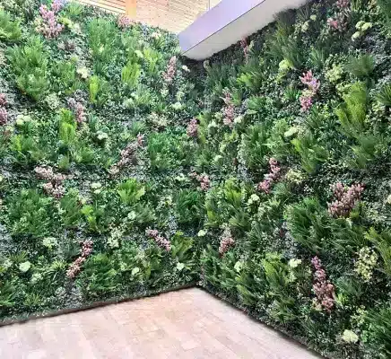 A room with walls covered in lush green foliage and pink flowers, creating a vertical garden effect through artificial design. The floor is beige wood, and the ceiling features natural light filtering in from above.