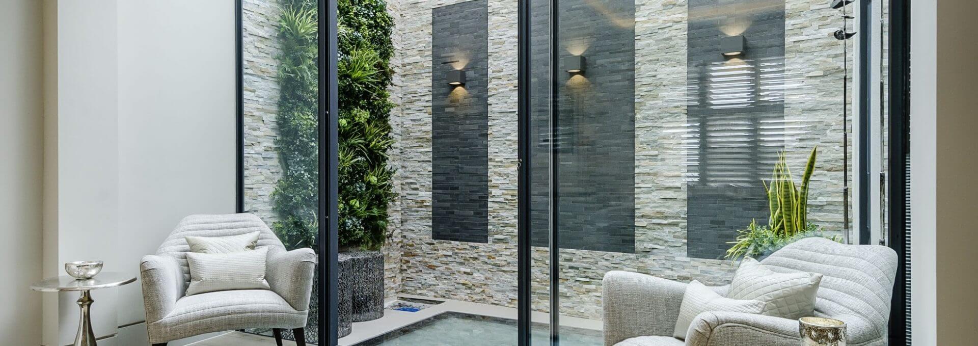 Modern indoor space with glass walls, featuring two white armchairs and a small side table. A custom-fit green wall is mounted on a stone backdrop, with subtle lighting and contemporary decor creating a serene atmosphere.