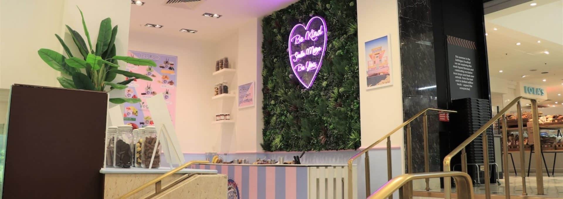 A cozy ice cream shop with a pastel color scheme features a neon sign reading Real Neat Ice Cream Cakes mounted on a leafy retail green wall, perfect for social media snaps. The counter displays assorted toppings in jars, while a potted plant adds greenery to the corner. Steps lead to the entrance.