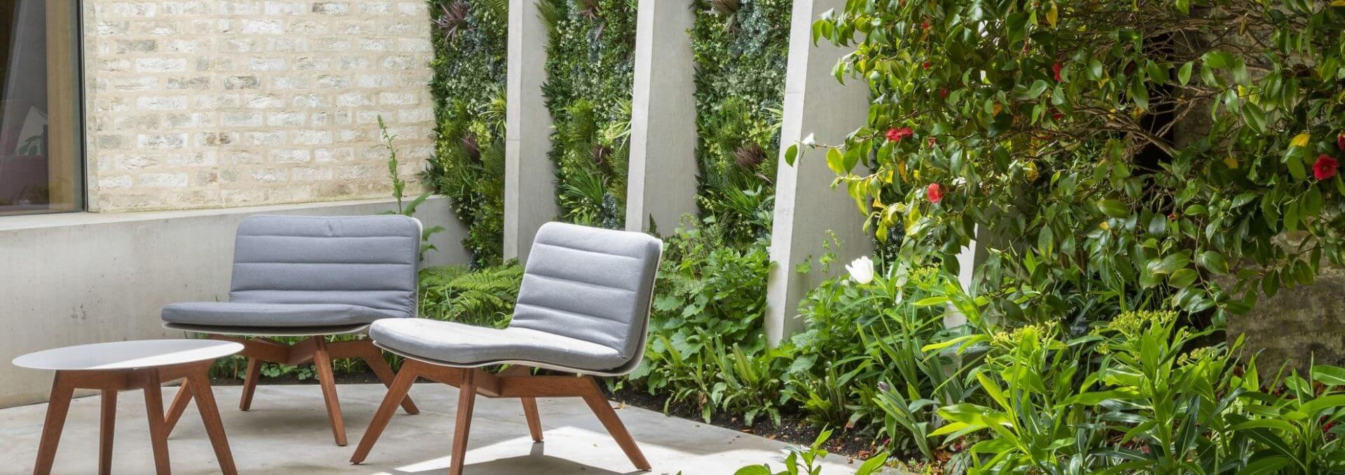 A garden patio boasts two modern gray chairs and a small white table set on concrete. Lush greenery, enhanced by an artificial green wall, surrounds the space, complemented by a brick backdrop. Vertical slats offer partial shade, creating a serene retreat.