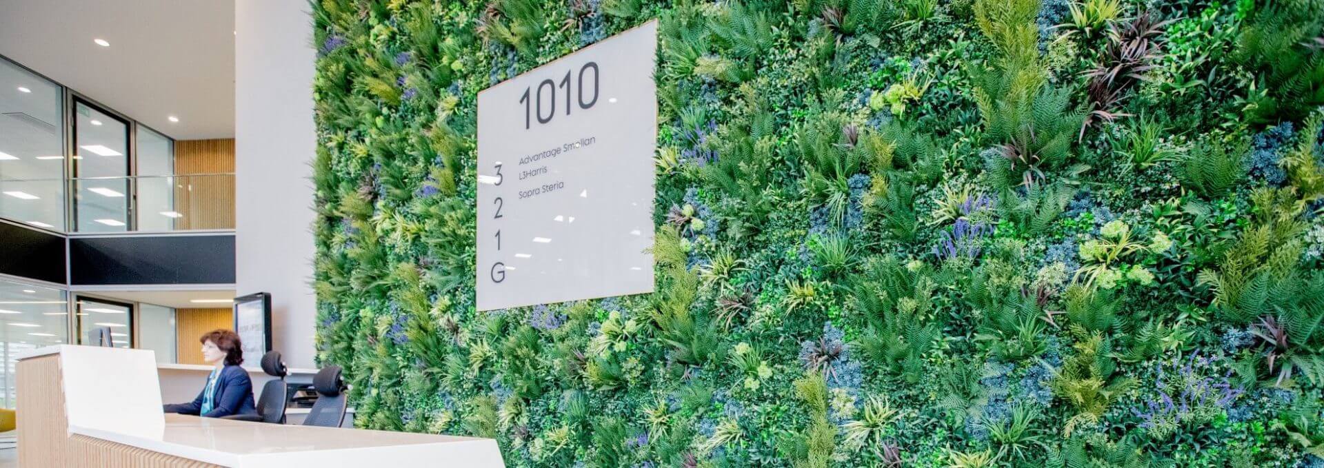 The modern lobby of the Green Wall Business Park in Reading features a lush vertical garden with diverse plants. A sign displays 1010, and a reception desk extends in the foreground. A person sits at the desk, with open office space visible to the left.