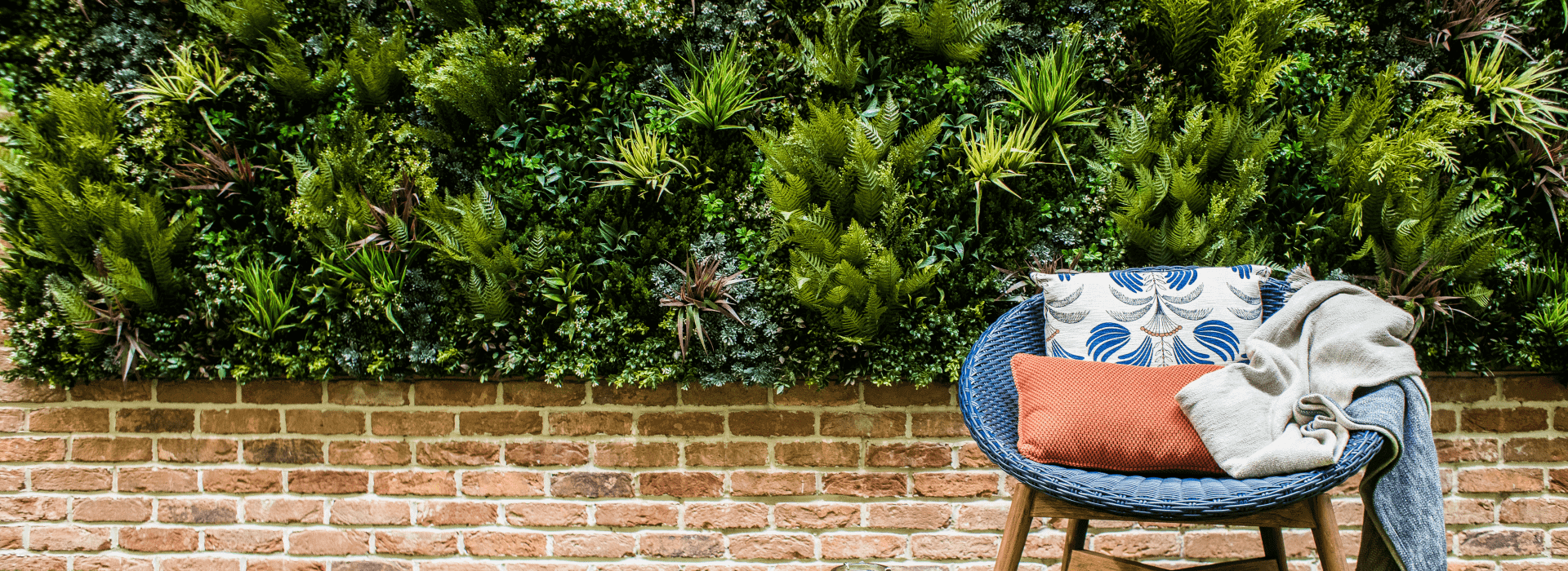 A blue wicker chair with a white and blue pillow, an orange pillow, and a gray blanket sits in front of a thriving green wall on the roof terrace. The wall beneath is made of red brick.