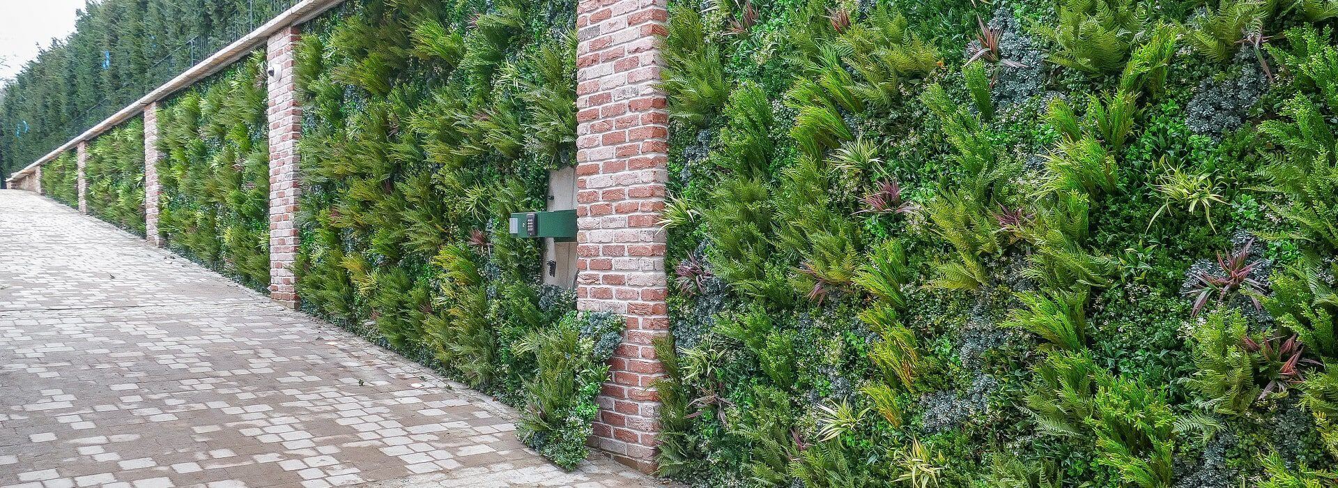 A brick wall with vertical green plants growing between the bricks creates a lush facade, embodying the natural-looking green wall often admired in Surrey, UK. A paved sidewalk runs alongside, bordered by trees.