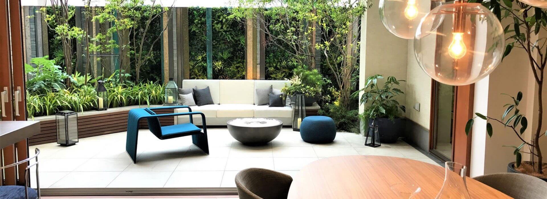 A modern living room opens to a lush garden patio, reminiscent of a serene green wall in Tokyo. It features a comfortable white sofa, a blue chair, and a circular table. Hanging globe lights and greenery complete the tranquil ambiance.