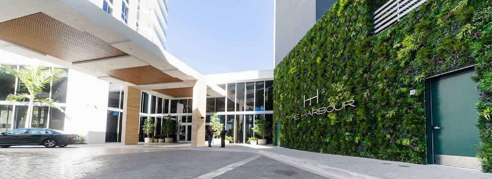 Outdoor view of a modern building complex with a lush large-scale green wall installation in Miami, Florida, displaying the words The Harbour. The entrance features wooden accents, large glass windows, and a parked car visible to the left. Bright, sunny day.