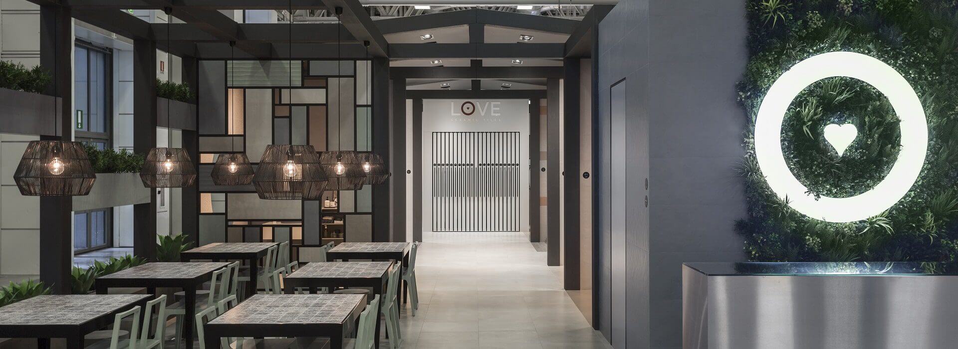 Contemporary indoor dining space in Portugal featuring hanging geometric pendant lights above tables. A wall with a heart-shaped light fixture and a private green wall in Lisbon is on the right. The background showcases a wall with the word LOVE.