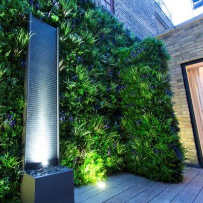 A Soft Lavender Colour Box wall adorned with lush greenery is illuminated at night. Nearby, a tall, modern fountain with a textured surface and spotlight stands on a wooden deck, casting light on the plants and creating a serene outdoor ambiance.