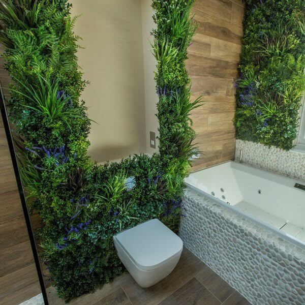 A modern bathroom featuring a wall-mounted toilet and a bathtub surrounded by lush vertical greenery. The walls are adorned with wooden panels and decorative stone tiles. The design creates a natural and relaxing atmosphere.