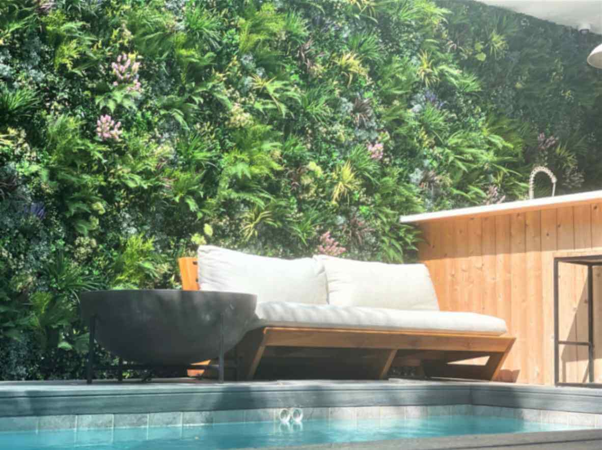 A cozy poolside scene features an artificial wooden lounge with white cushions beside a corner bar. The backdrop is a lush vertical garden with various green plants, creating a serene and inviting atmosphere.