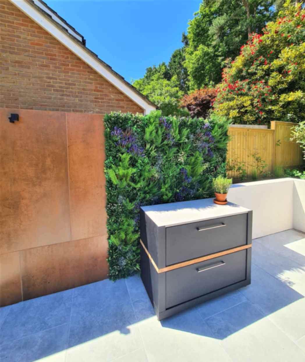 A modern outdoor patio with a small black cabinet featuring two drawers and a potted plant on top. The green wall is enhanced with vibrant plants, ideal for those wondering, "Can I install an artificial green wall outside?" A brick house and wooden fence complete the sunny scene.