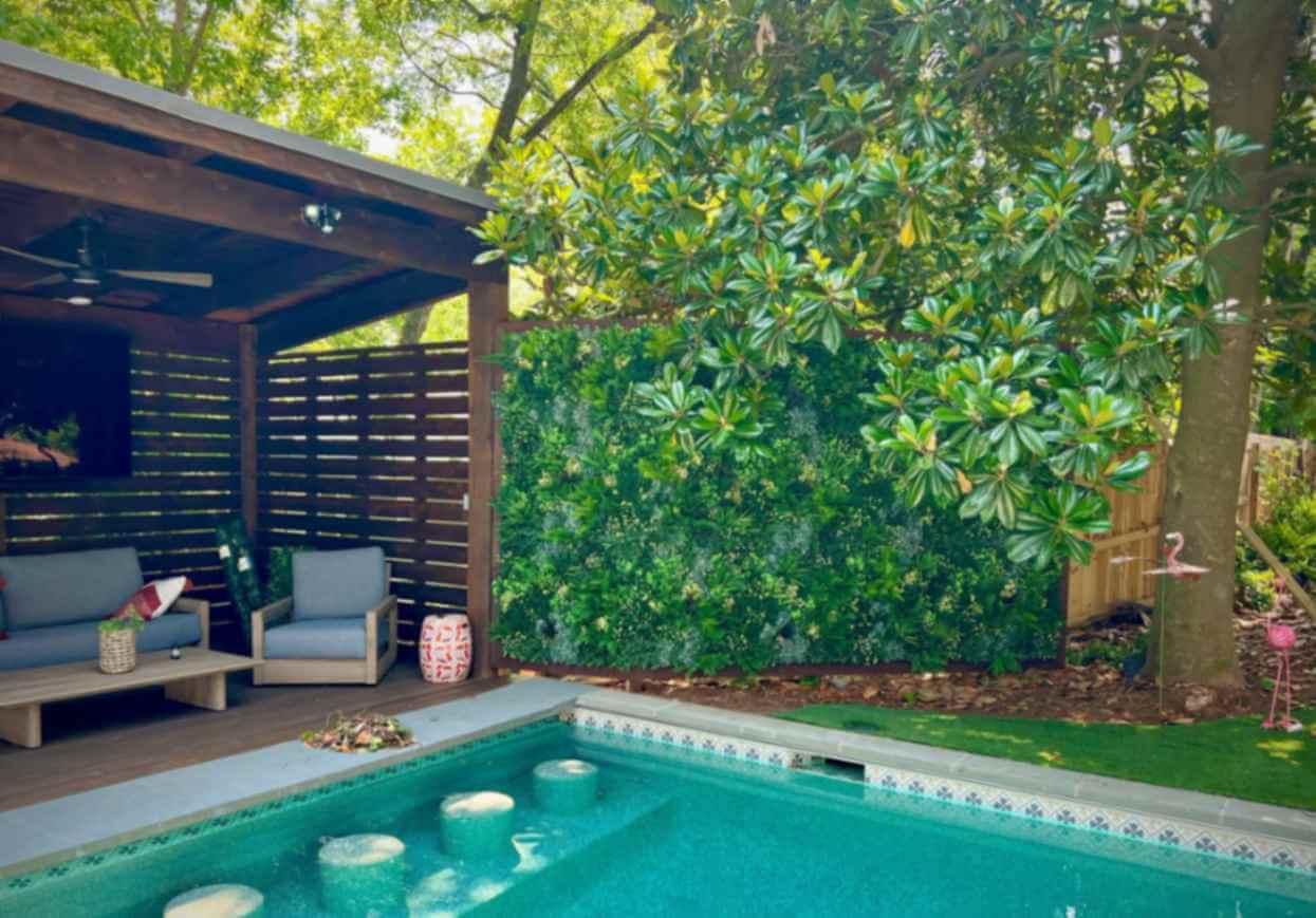 A backyard setup in Atlanta, Georgia, boasts a swimming pool with bar stools in the water. To the left, there's a covered seating area with gray chairs and a ceiling fan. Lush green foliage and wooden backyard privacy screening border the area, accompanied by playful flamingo lawn ornaments.