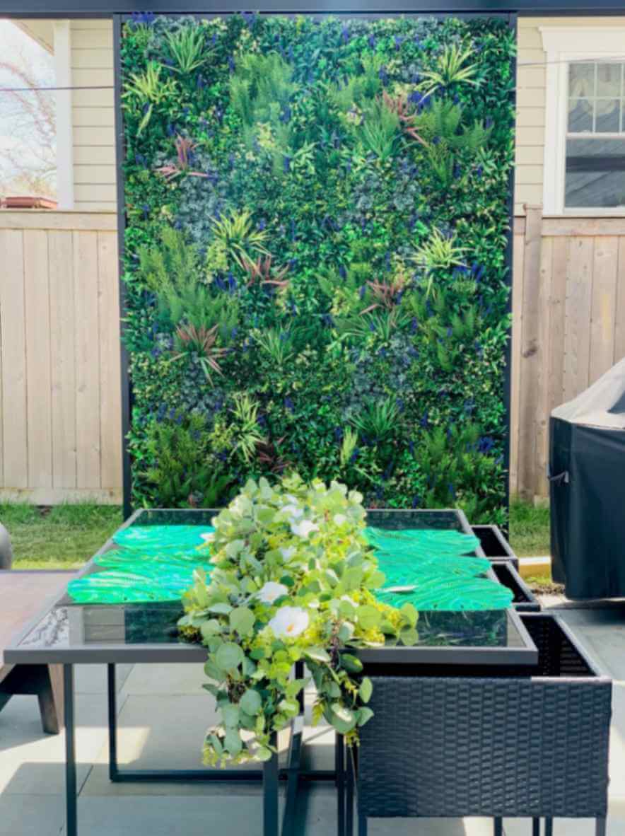 A patio scene features a black outdoor dining table with a lush green runner centerpiece. In this backyard, a vibrant green wall offers an Illinois touch, displaying various plants that add natural charm to the setting. A wooden fence completes the backdrop, perfect for a Chicago oasis.