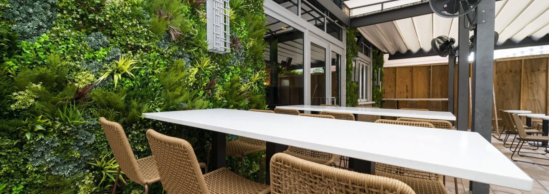 Outdoor seating area with white tables and wicker chairs. A lush green wall provides a touch of cafe privacy in Miami's vibrant atmosphere. The space, covered by a pergola with a fan, is enclosed by glass doors on one side, creating an inviting oasis perfect for Florida’s sunny days.