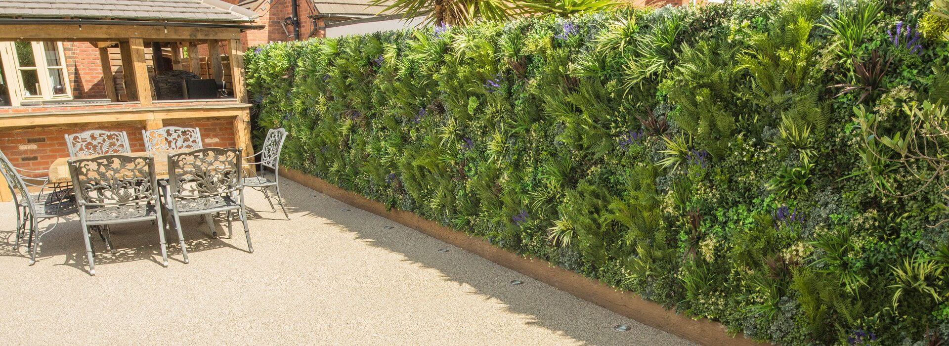 A patio with ornate metal chairs and tables is next to a lush vertical garden wall. The wall is densely covered with various green plants, creating a vibrant natural backdrop. A brick building and roof are visible in the background.