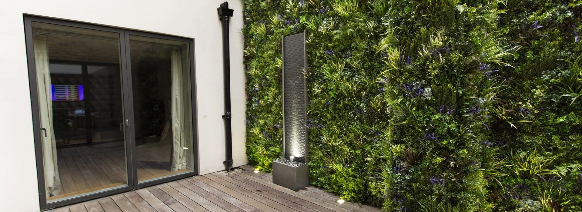 A modern patio with a glass sliding door on the left and an artificial green wall on the right, featuring a tall, decorative water feature in front of it. The floor is made of wooden planks, creating a serene outdoor space.