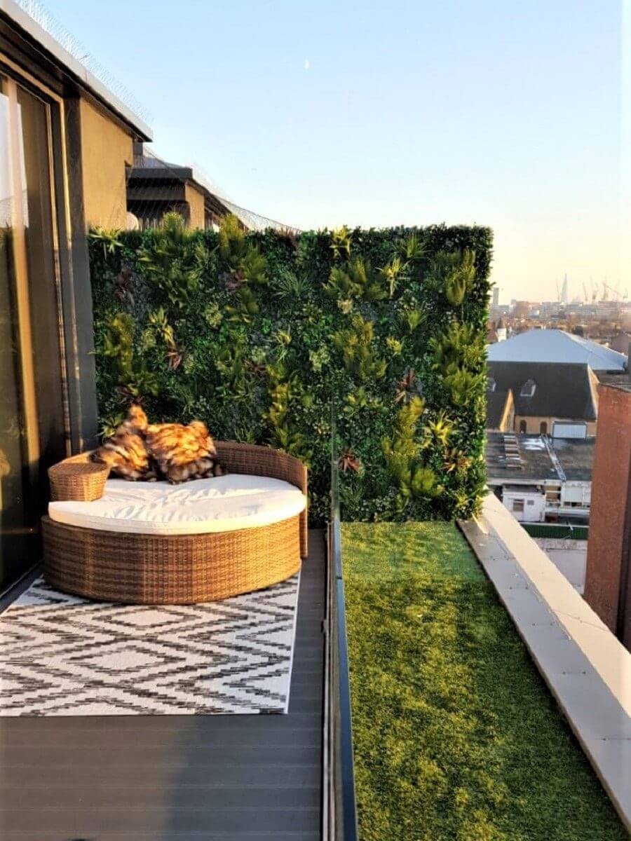 A rooftop balcony features a round, wicker lounge chair with plush cushions, a vibrant green wall of realistic artificial plants, a patterned rug, and soft artificial grass. The view overlooks the cityscape under a clear sky.