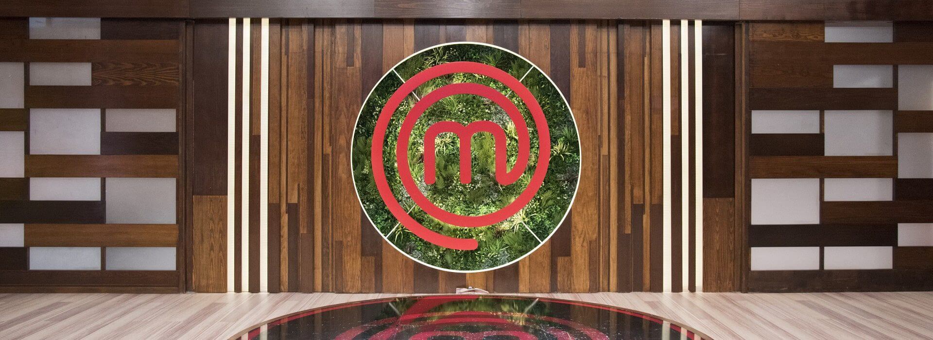 A modern, wooden wall boasts a large circular emblem with a red swirl design reminiscent of the Masterchef logo, set against a lush green wall backdrop. The floor reflects this striking emblem, adding to the sleek and contemporary design perfect for TV settings.