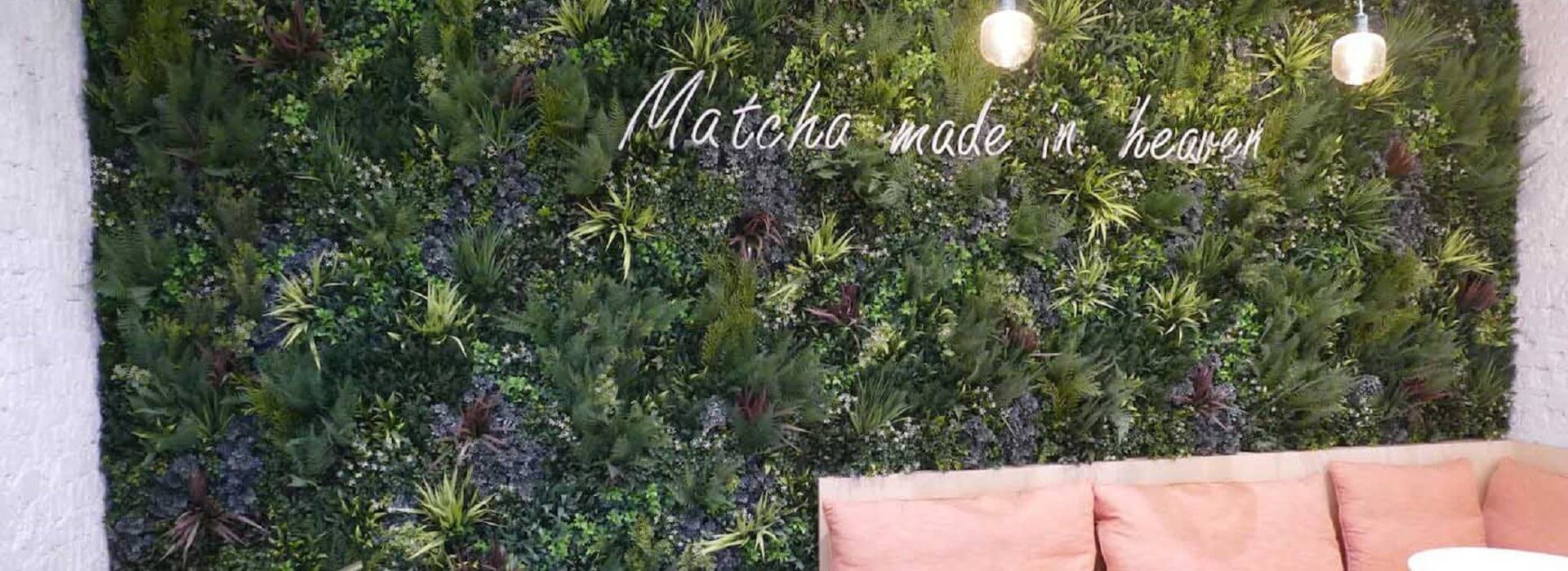 A vertical garden wall with lush green plants and two ceiling lights complements the coffee shop's design in London, UK. The wall features neon text reading "Matcha made in heaven." Below, pink cushions rest on a light bench, inviting you to relax and enjoy the atmosphere.