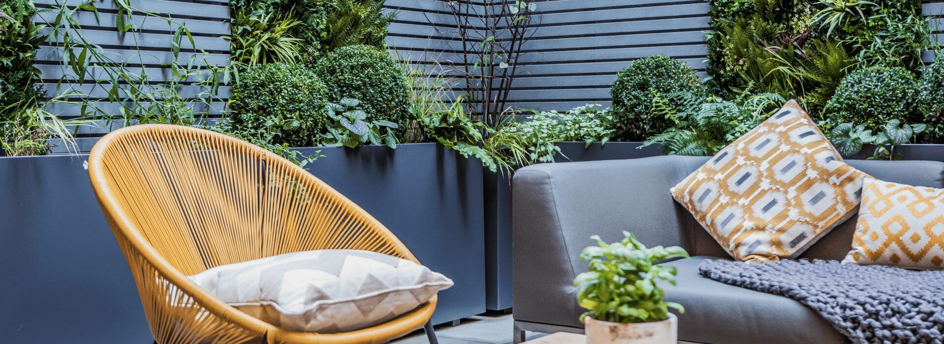 Modern outdoor patio with a yellow woven chair, gray sofa adorned with patterned cushions, and a small potted plant on a wooden table. Enhanced by the courtyard faux green wall design, lush plants stand tall in planters against the slatted gray wall, creating an urban oasis in London, UK.