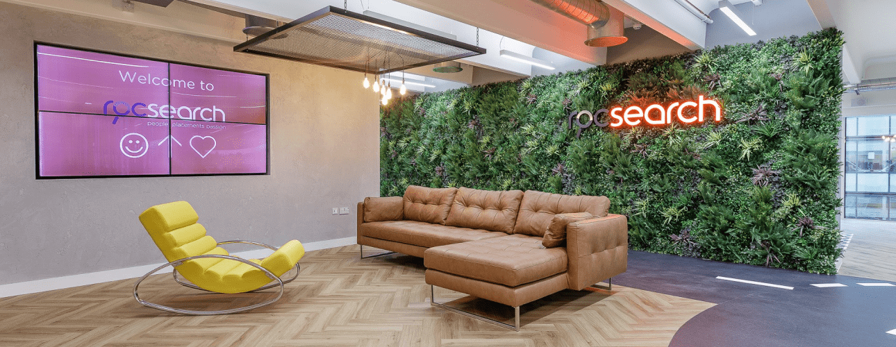 The office reception area showcases a large green wall with a neon presence sign, conveying an artificial charm. A brown sofa and a yellow rocking chair rest on the herringbone floor. On the left wall, a screen displays a welcome message, while ceiling lights add a modern touch.