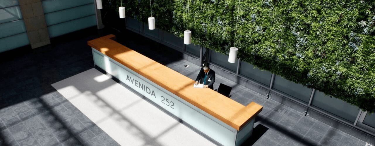 A large, modern reception desk labeled AVENIDA 252 is positioned in an open space with a person seated behind it. The area features a vertical green wall with plants and several hanging pendant lights, showcasing our key advantage of having office screening while sunlight streams through tall windows.