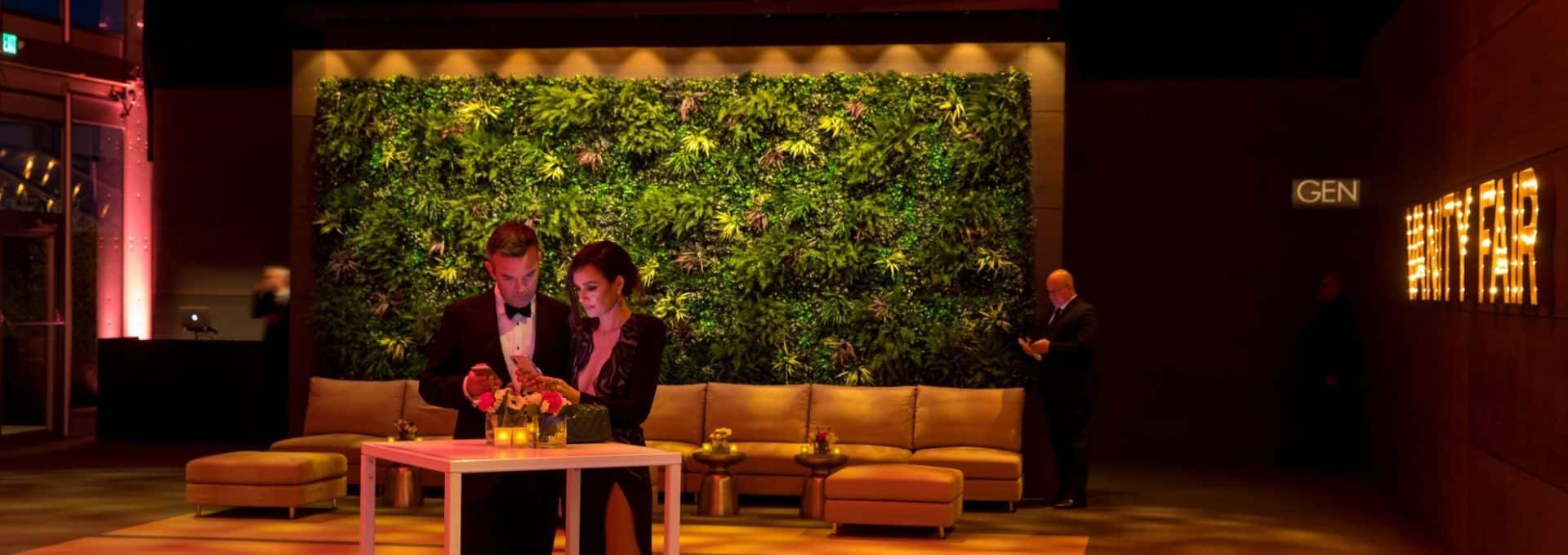 In the dimly lit lounge of the Vanity Fair Oscars Party Green Wall in Beverly Hills, two people in formal attire sit at a small table, intently looking at a phone. A lush green wall of plants and a man in the background are visible, with the word GEN partially illuminated on a wall.